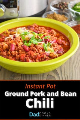 A bowl of ground pork and bean chili, sprinkled with cheese and chopped green onion, with spices, sliced jalapeños, and an Instant Pot in the background