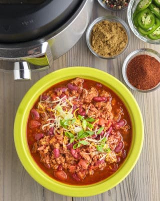 A bowl of ground pork and bean chili, sprinkled with cheese and chopped green onion, with spices, sliced jalapeños, and an Instant Pot in the background