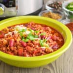 A bowl of ground pork and bean chili, sprinkled with cheese and chopped green onion, with spices, sliced jalapeños, and an Instant Pot in the background