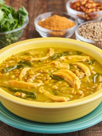 A yellow bowl of rotisserie chicken and lentil soup, with spices and herbs in the background