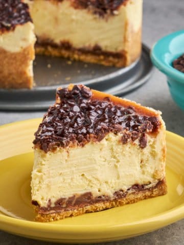 A slice of chocolate chip cheesecake on a yellow plate, with a bowl of mini chocolate chips and the cheesecake in the background