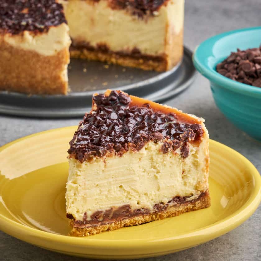A slice of chocolate chip cheesecake on a yellow plate, with a bowl of mini chocolate chips and the cheesecake in the background