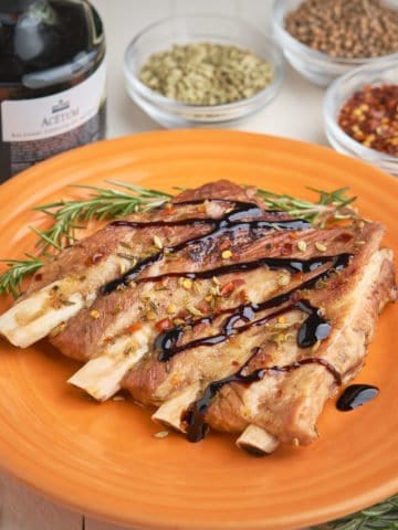 A slab of 4 ribs, sprinkled with herbs and drizzled with balsamic glaze, on an orange plate, with herbs and balsamic vinegar in the background.