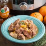 A blue bowl of braised pork, with orange zest and rosemary sprinkled on top, and an Instant Pot, oranges, honey bear, and rosemary sprigs in the background
