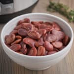 A white bowl of Christmas lima beans on a wood table with an Instant Pot and sprig of thyme in the background.