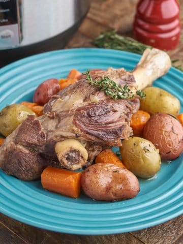 A braised lamb shank, surrounded by potatoes and carrots, on a teal plate, with an Instant Pot, a bundle of thyme, and a pepper mill in the background