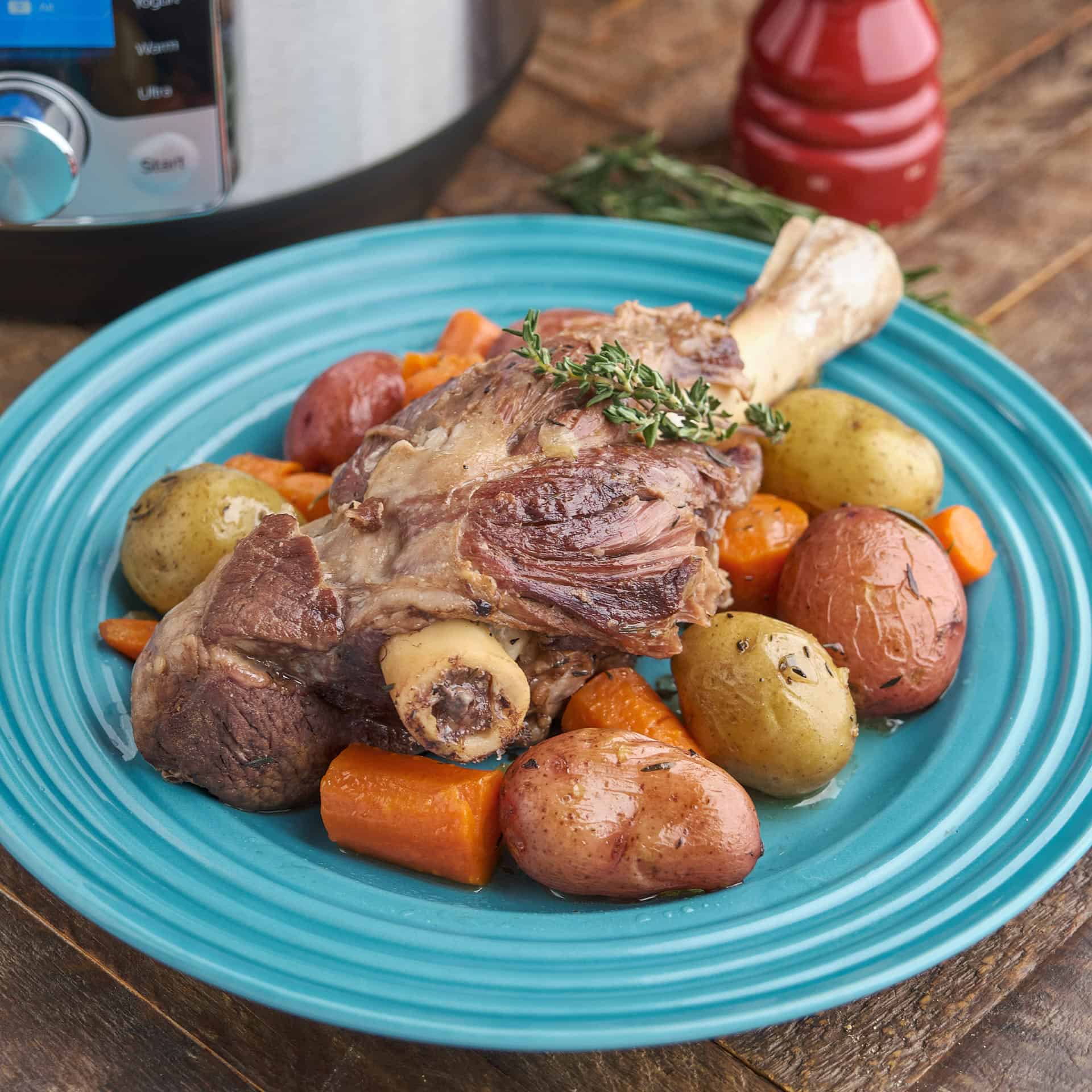 A braised lamb shank, surrounded by potatoes and carrots, on a teal plate, with an Instant Pot, a bundle of thyme, and a pepper mill in the background