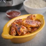 Gochujang glazed chicken wings sprinkled with sesame seeds in a yellow dish, with a bowl of gochujang, a bowl of rice, and an Instant Pot in the background.