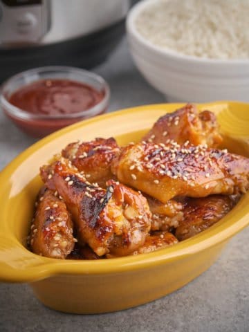 Gochujang glazed chicken wings sprinkled with sesame seeds in a yellow dish, with a bowl of gochujang, a bowl of rice, and an Instant Pot in the background.