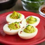 Four pesto deviled eggs, with green filling, sprinkled with basil and red pepper flakes, on a maroon plate