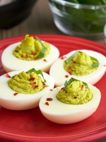 Four pesto deviled eggs, with green filling, sprinkled with basil and red pepper flakes, on a maroon plate