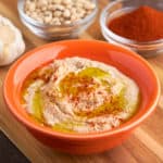 A bowl of black-eyed pea hummus, drizzled with olive oil and sprinkled with paprika, with a bowl of paprika, black-eyed peas, and garlic in the background