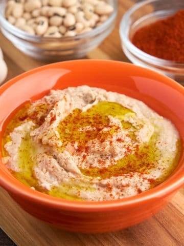 A bowl of black-eyed pea hummus, drizzled with olive oil and sprinkled with paprika, with a bowl of paprika, black-eyed peas, and garlic in the background