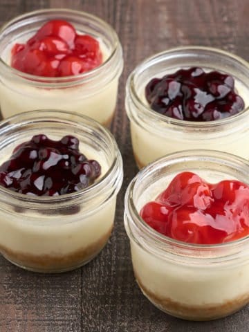 Four mini cheesecakes in canning jars, with cherry and blueberry topping, on a wood tabletop