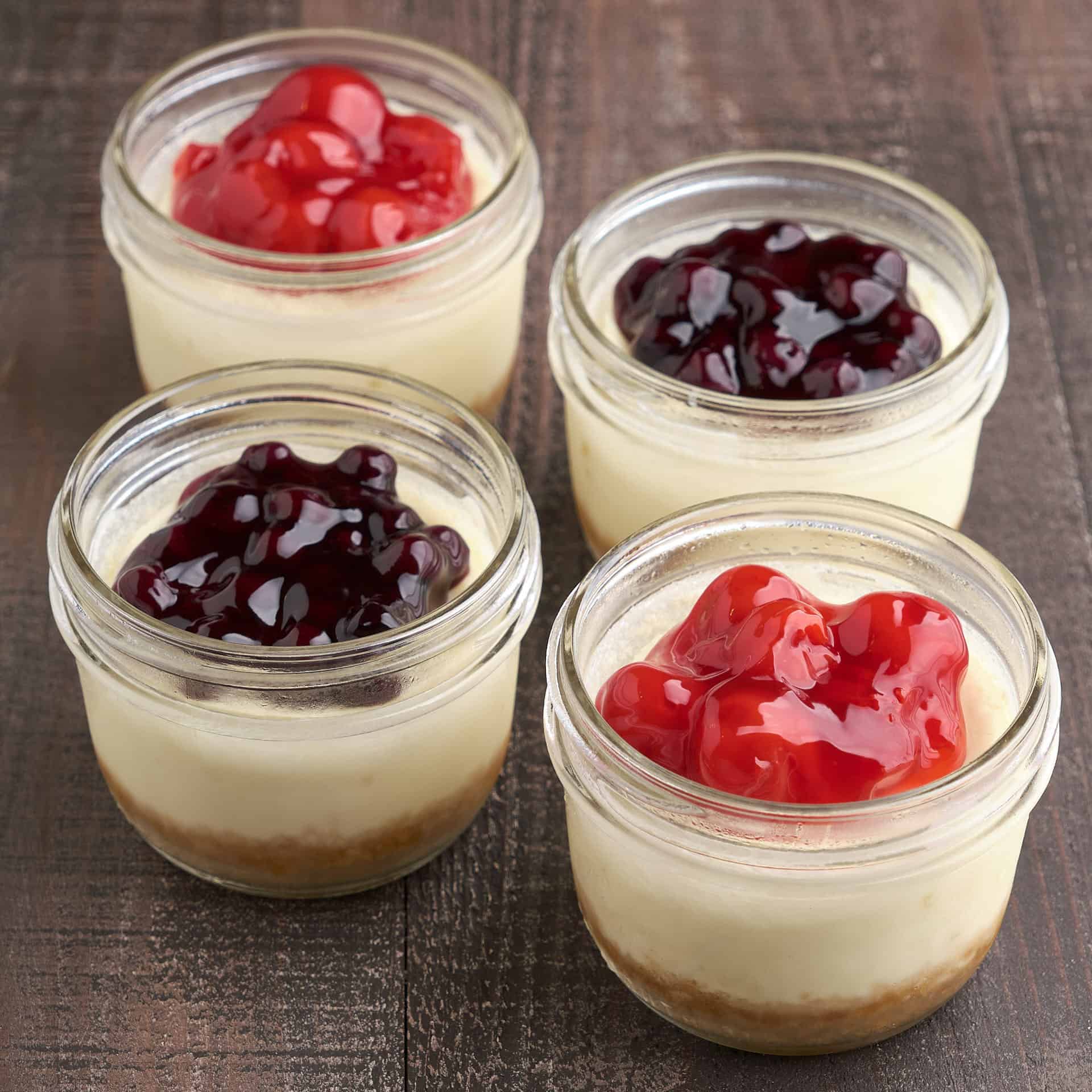 Four mini cheesecakes in canning jars, with cherry and blueberry topping, on a wood tabletop