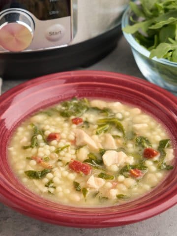 A bowl of chicken and herb soup, with bits of chicken, pancetta, herbs, and pasta visible, and an Instant Pot and bowl of arugula in the background.