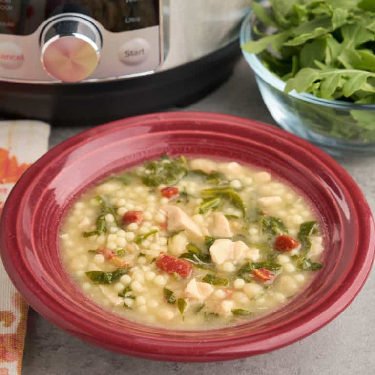 A bowl of chicken and herb soup, with bits of chicken, pancetta, herbs, and pasta visible, and an Instant Pot and bowl of arugula in the background.