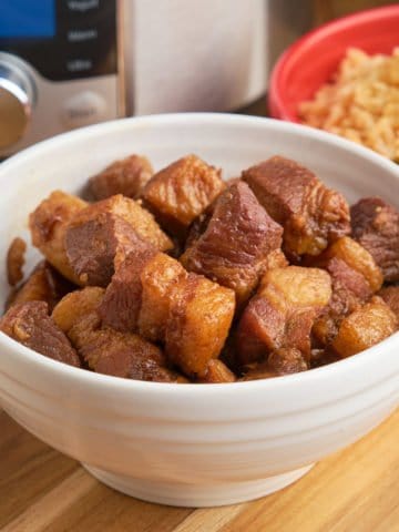 A bowl of red braised pork belly cubes in front of an Instant Pot and a bowl of rice