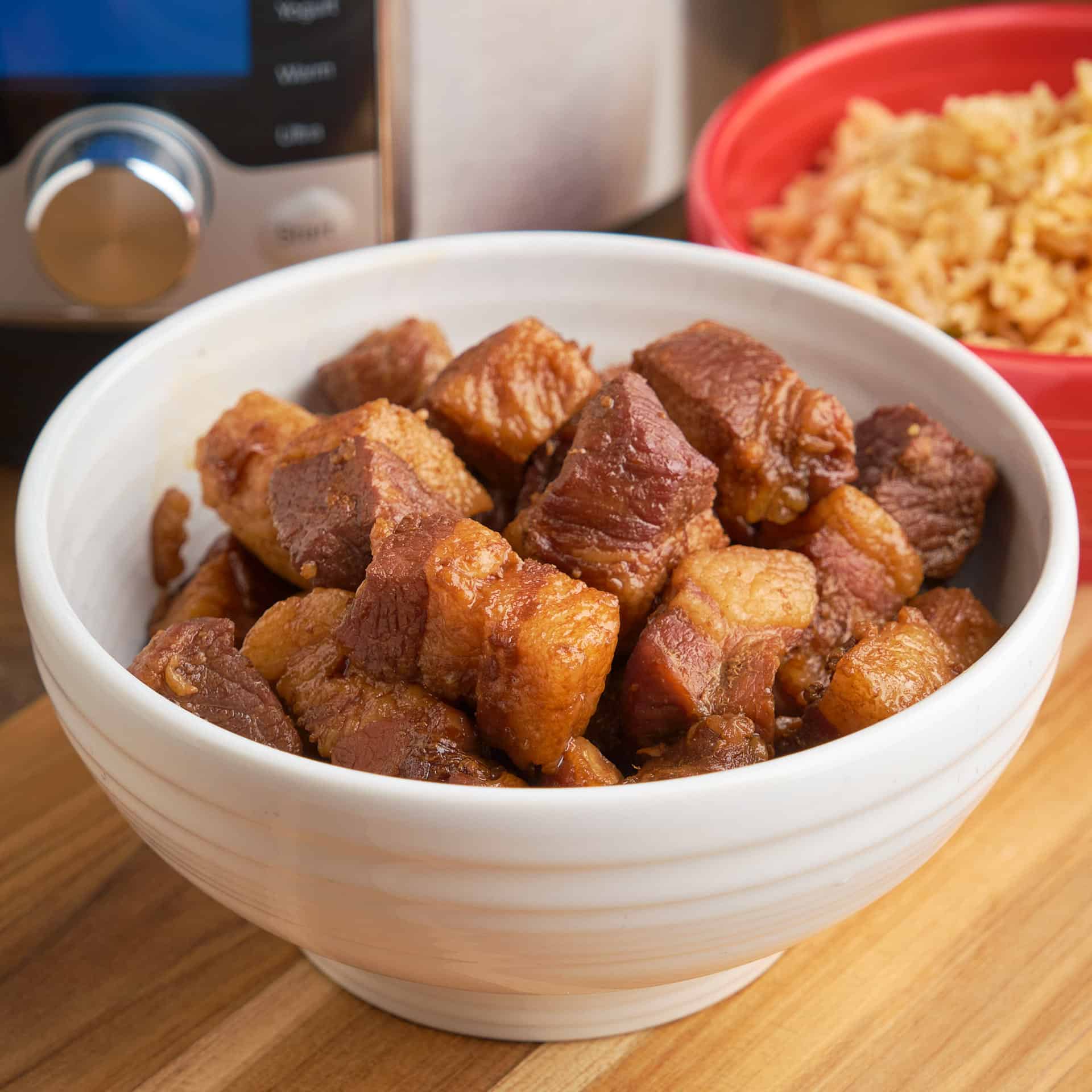 A bowl of red braised pork belly cubes in front of an Instant Pot and a bowl of rice