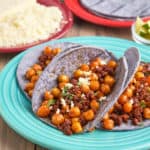 Chickpea and chorizo in blue corn tortillas, sprinkled with crumbled queso fresco and diced cilantro, with more queso, blue corn tortillas, and lime wedges in the background.