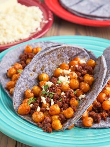 Chickpea and chorizo in blue corn tortillas, sprinkled with crumbled queso fresco and diced cilantro, with more queso, blue corn tortillas, and lime wedges in the background.