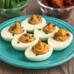 Sun-dried tomato deviled eggs on a plate, sprinkled with basil, with basil leaves and sun-dried tomatoes in the background.