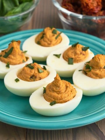 Sun-dried tomato deviled eggs on a plate, sprinkled with basil, with basil leaves and sun-dried tomatoes in the background.