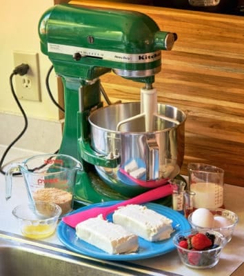 A green stand mixer in front of plates, bowls, and measuring cups holding the ingredients for berry cheesecake