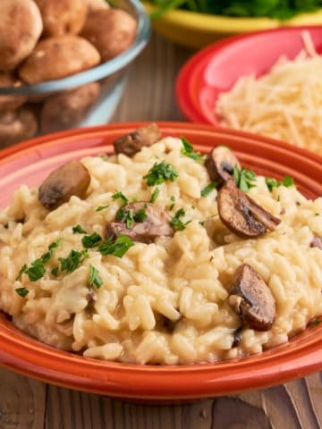 A bowl of mushroom risotto, with mushrooms, cheese, and parsley in the background