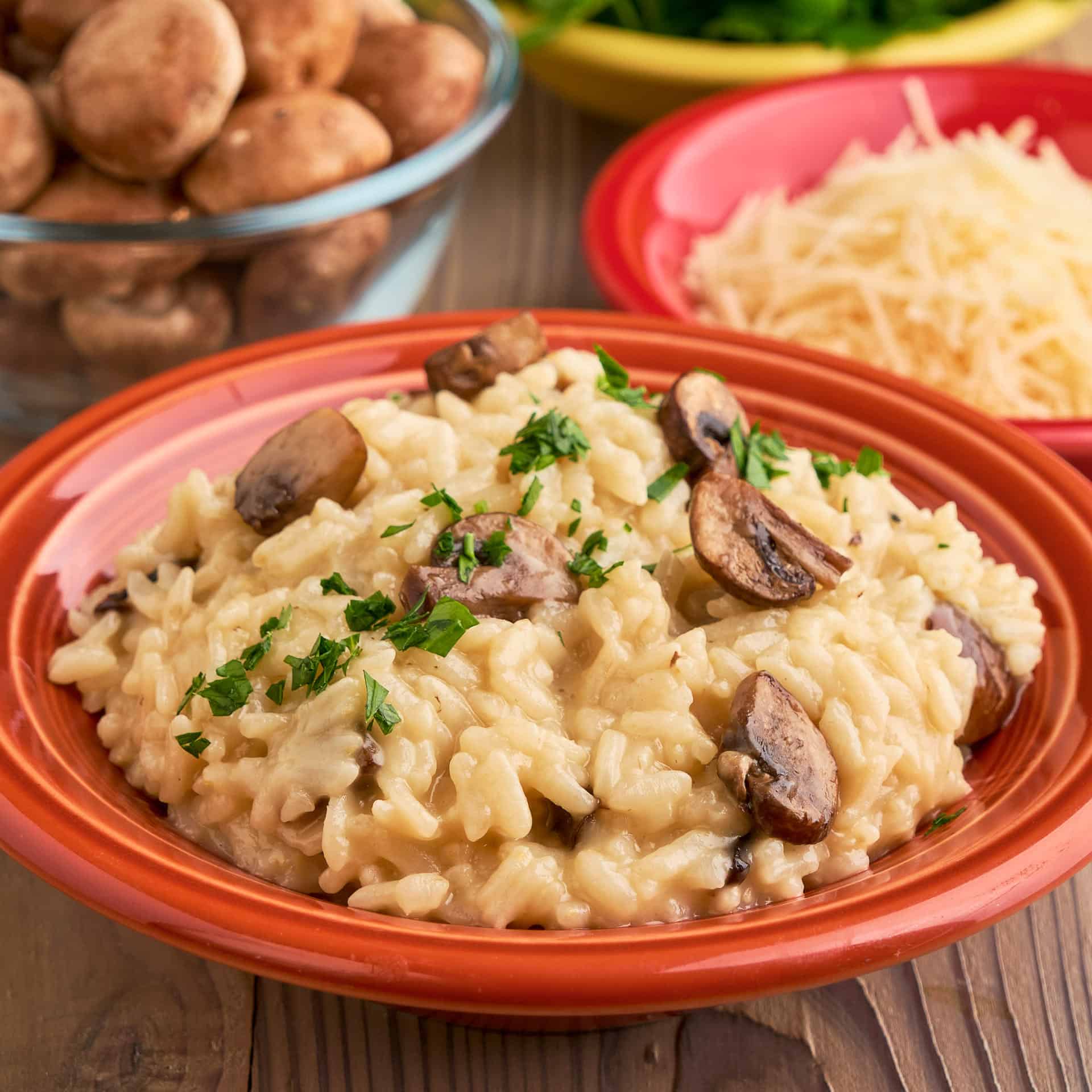 A bowl of mushroom risotto, with mushrooms, cheese, and parsley in the background