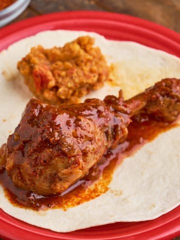 An Ethiopian chicken drumstick, coated with sauce, on a tortilla on a red plate