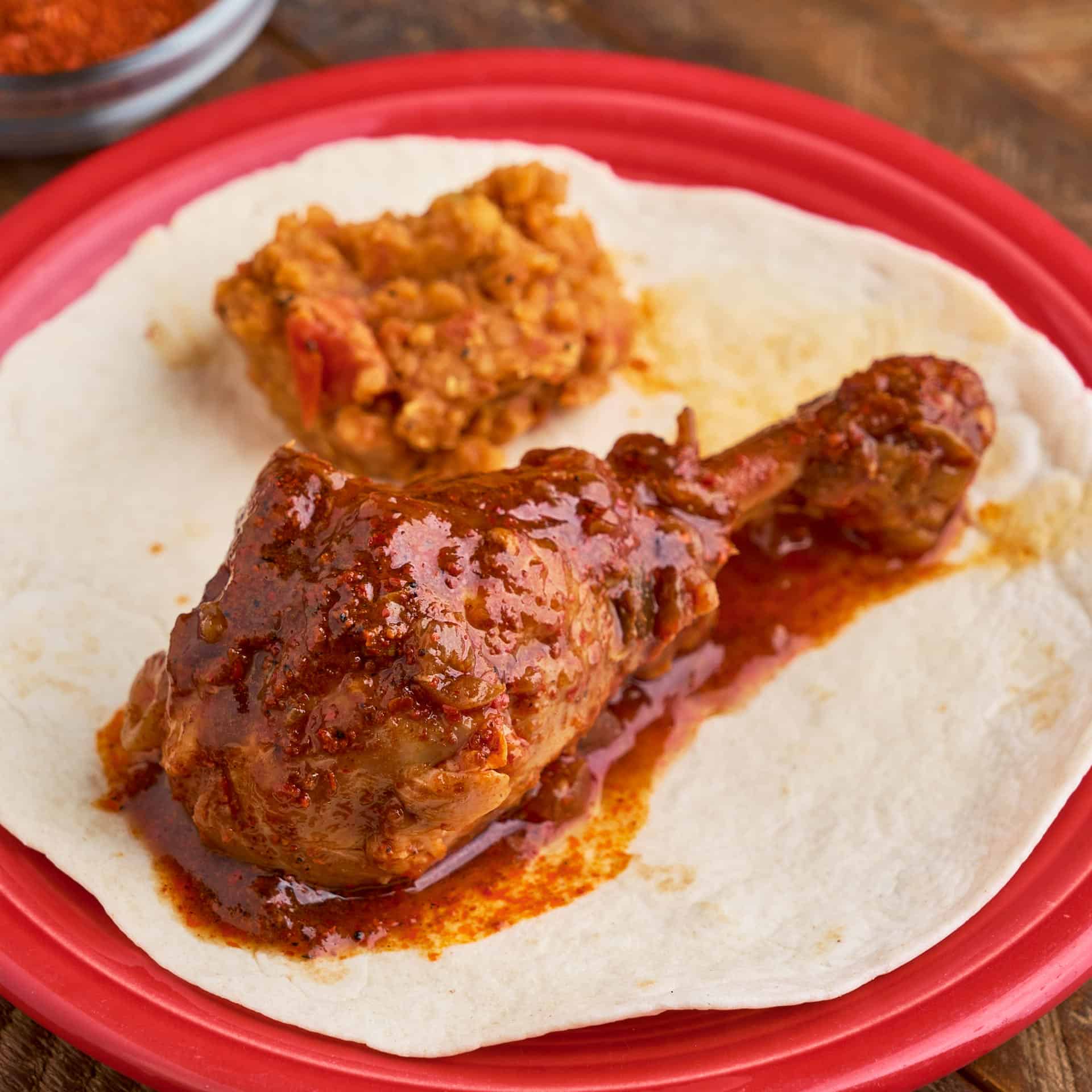 An Ethiopian chicken drumstick, coated with sauce, on a tortilla on a red plate