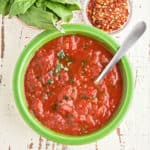 A bowl of arrabbiata sauce sprinkled with slivered basil, and a spoon sticking out of it, with a bowl of pepper flakes and a bowl of basil.