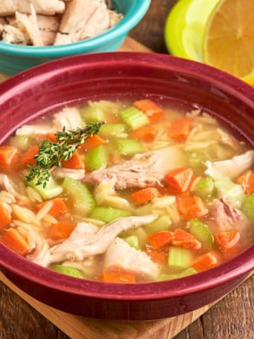 A bowl of lemon turkey and orzo soup, with a sprig of thyme, and a lemon and shredded turkey in the background