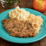 A pork shoulder chop on a teal plate with mashed potatoes and sauerkraut, with an apple and thyme in the background