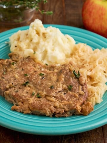 A pork shoulder chop on a teal plate with mashed potatoes and sauerkraut, with an apple and thyme in the background