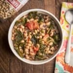 A bowl of black eyed peas and collard greens on a wood table with a bag of beans, a napkin, and a spoon