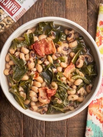 A bowl of black eyed peas and collard greens on a wood table with a bag of beans, a napkin, and a spoon