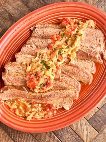 A platter of sliced brisket, covered with onions and tomatoes, on a wood table