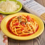 A plate of Bucatini all'Amatriciana, with a bowl of cheese and a bag of dried bucatini in the background