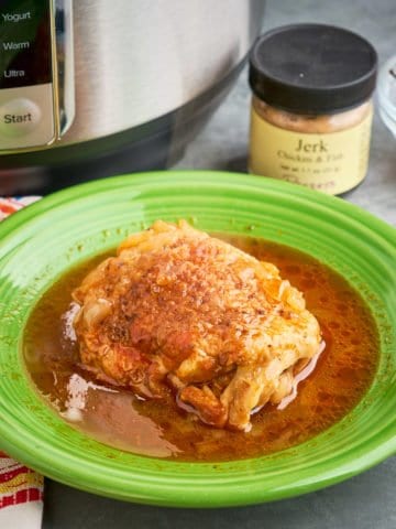 A chicken thigh in Jamaican jerk chicken fricassee sauce, on a green plate, in front of an Instant Pot, a jar of Jerk seasoning, and a Scotch Bonnet pepper