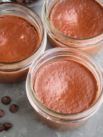 Three bittersweet chocolate pots de creme, with some chocolate chips on a gray tbable