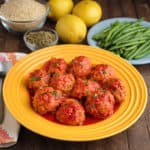 A plate of Greek meatballs in tomato sauce, with green beans, lemons, oregano, and rice in the background