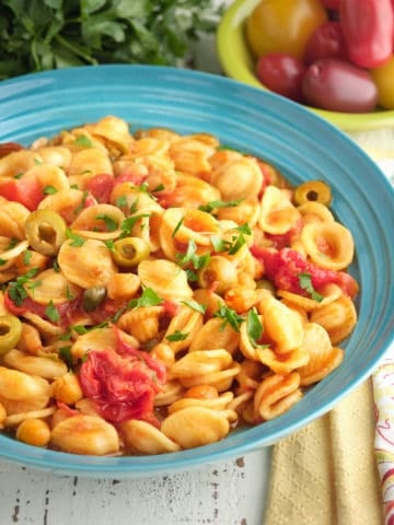 A bowl of chickpea puttanesca with orecchiette, with tomatoes, parsley, napkins, and a spoon