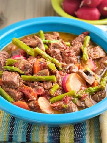 A bowl of beef stew with asparagus, carrots, and radishes.