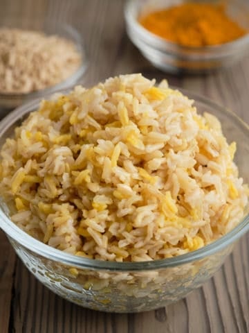 A bowl of brown basmati rice with uncooked rice and ground turmeric in the background