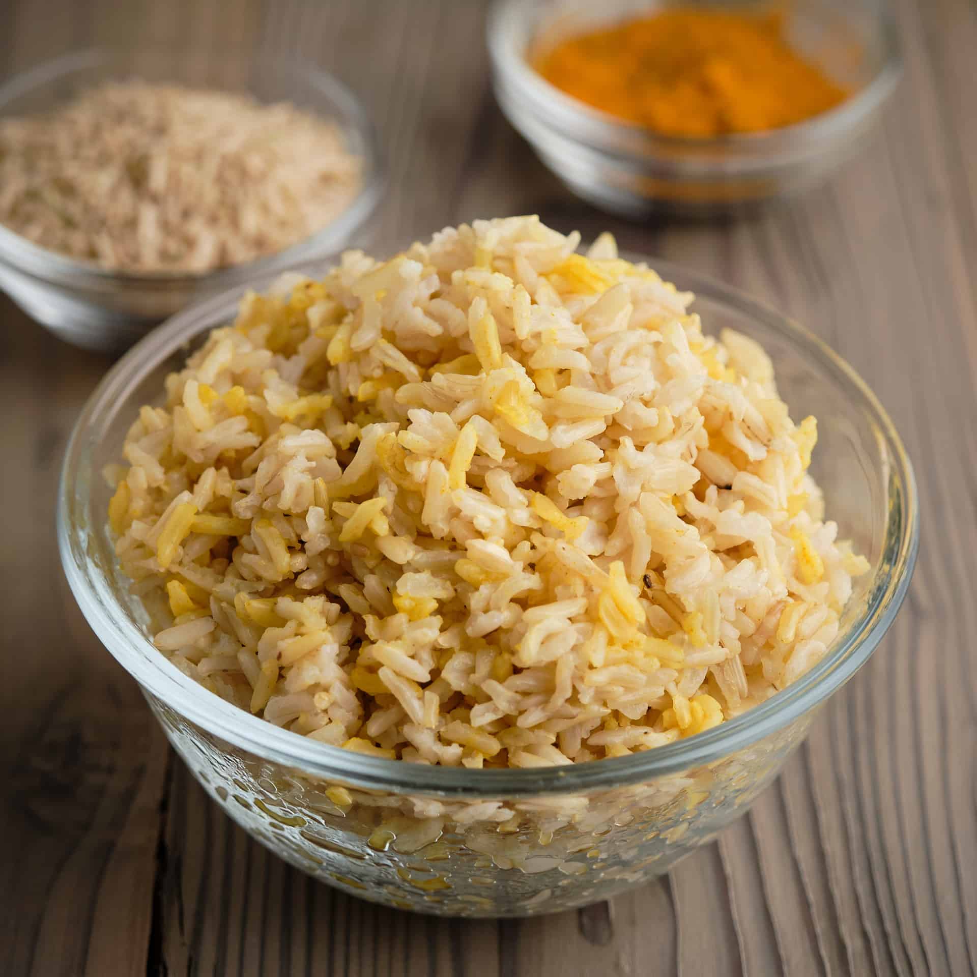 A bowl of brown basmati rice with uncooked rice and ground turmeric in the background