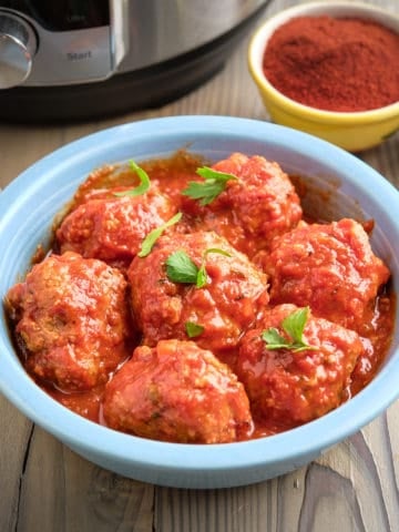 A bowl of Spanish meatballs (Albondigas) in tomato sauce, with a pressure cooker and smoked paprika in the background.