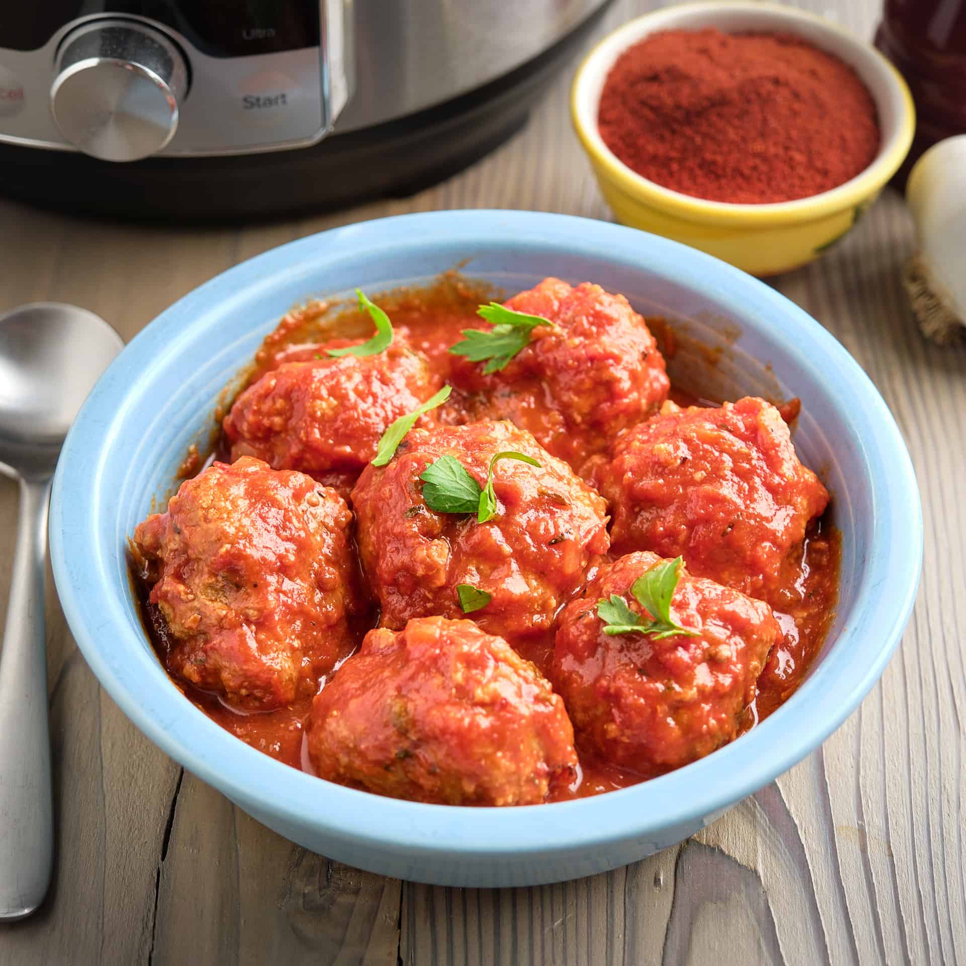 A bowl of Spanish meatballs (Albondigas) in tomato sauce, with a pressure cooker and smoked paprika in the background.