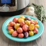 A plate of rainbow baby potatoes sprinkled with garlic and rosemary, with a sprig of rosemary, a head of garlic, and a pressure cooker in the background